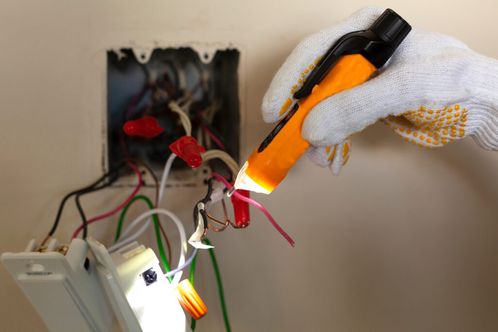 An electrician is repairing a wall switch. A DIY project concept. High voltage danger. A touchless voltage tester and protective rubber gloves are used. The tester has torch light for illumination for electrical repair